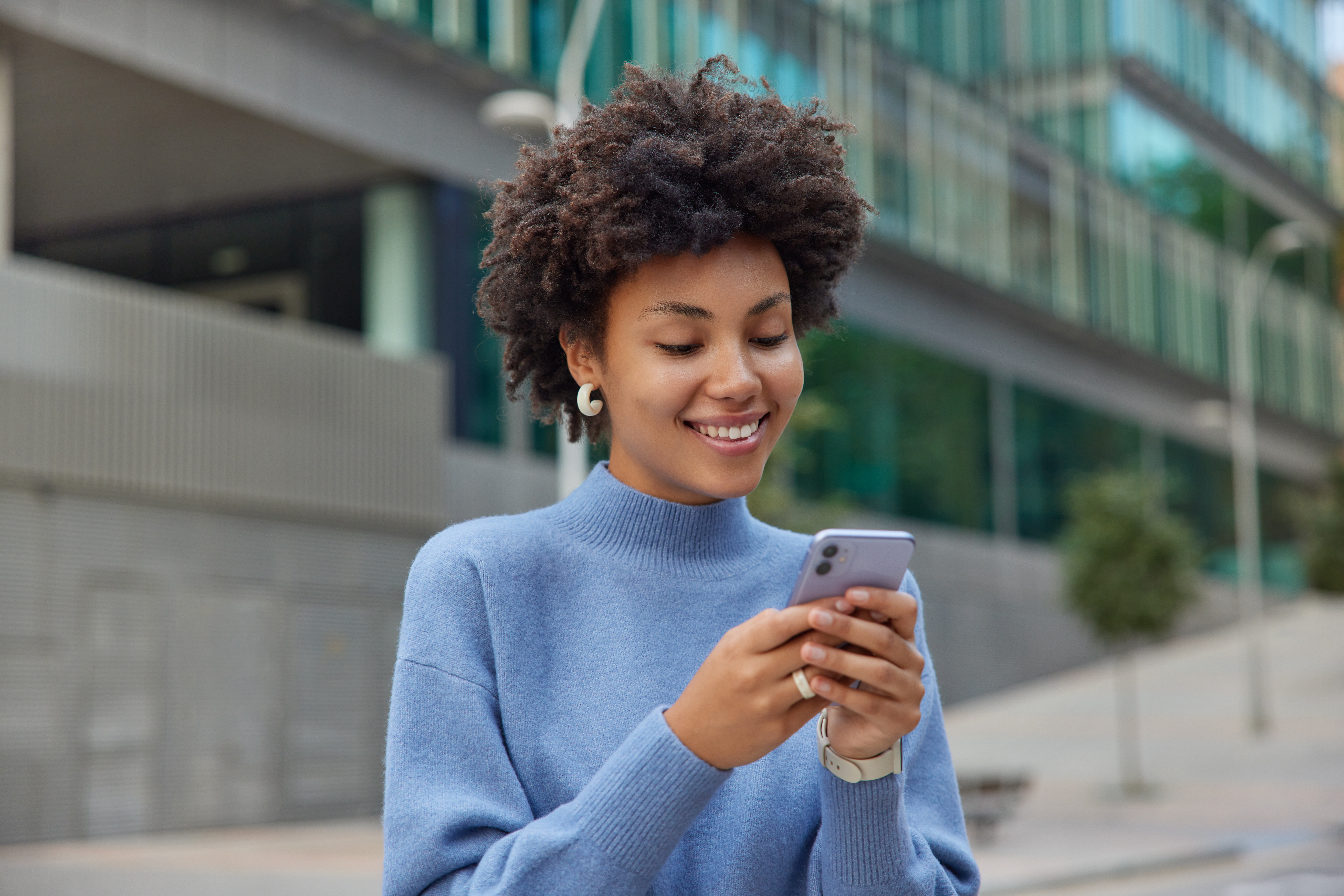 Glad young woman with curly bushy hair holds modern mobile phone downloads amazing application surfs social networks wears casual blue jumper smiles positively poses against blurred background