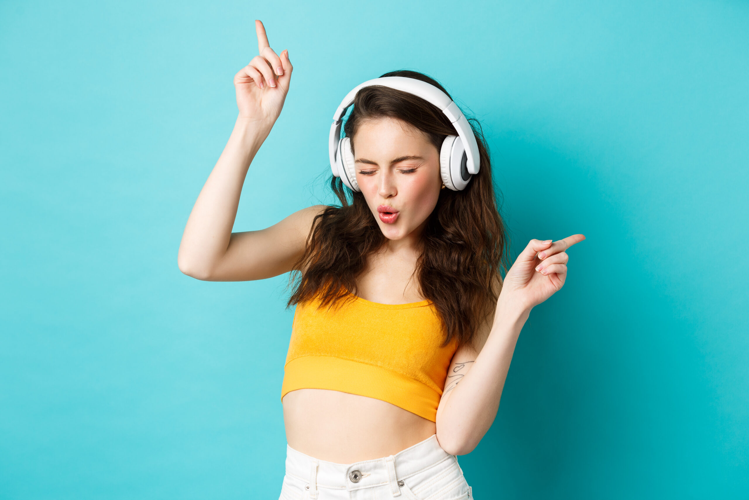 Stylish modern woman in headphones, listening music and dancing, enjoying favorite songs in earphones, standing against blue background
